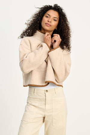 A woman with curly hair models the KELLY LUXE SHERPA REVERSIBLE JACKET by Velvet by Graham & Spencer, styled in beige over a white shirt and beige pants, posing against a plain light background.