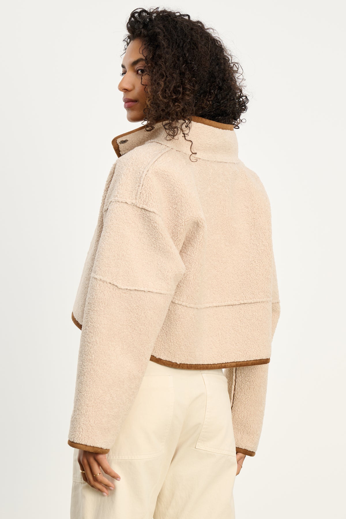 A person with curly hair wears the KELLY LUXE SHERPA REVERSIBLE JACKET by Velvet by Graham & Spencer, a beige cropped faux sherpa fleece with brown trim, seen from the back against a plain light background.-39838356013249