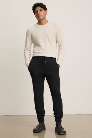 A person stands against a plain background wearing the Velvet by Graham & Spencer CROSBY JOGGER, a cream long-sleeve sweater, and black boots, with hands in pockets.