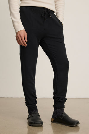 A person wears a cream sweater, Velvet by Graham & Spencer CROSBY JOGGER luxe fleece pants, dark socks, and black slip-on clogs, standing on a polished floor against a plain background.