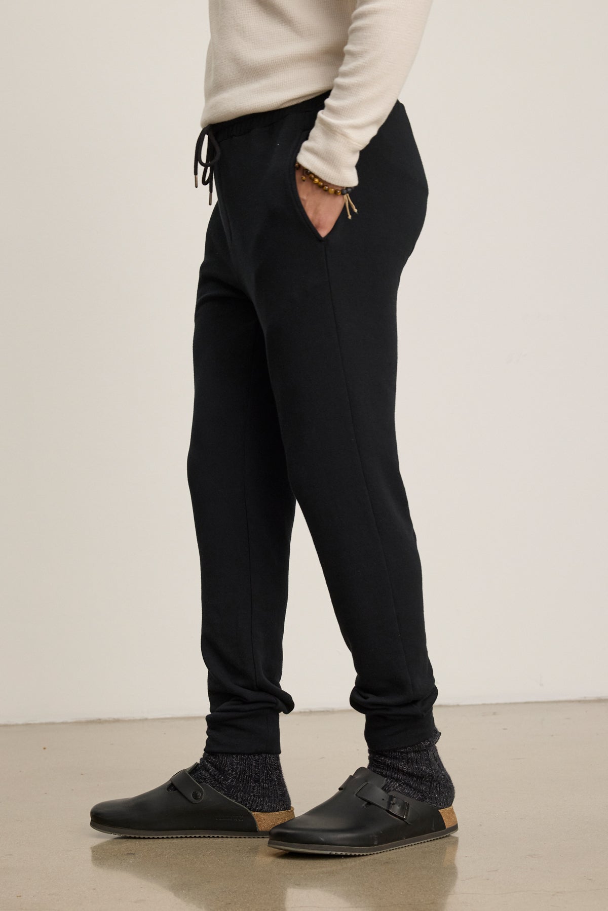 On a polished floor, someone wears Velvet by Graham & Spencer CROSBY JOGGER pants with a cream long-sleeve shirt, dark socks, and black slip-on shoes featuring tan soles.
