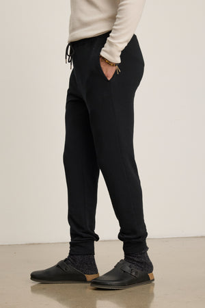 On a polished floor, someone wears Velvet by Graham & Spencer CROSBY JOGGER pants with a cream long-sleeve shirt, dark socks, and black slip-on shoes featuring tan soles.