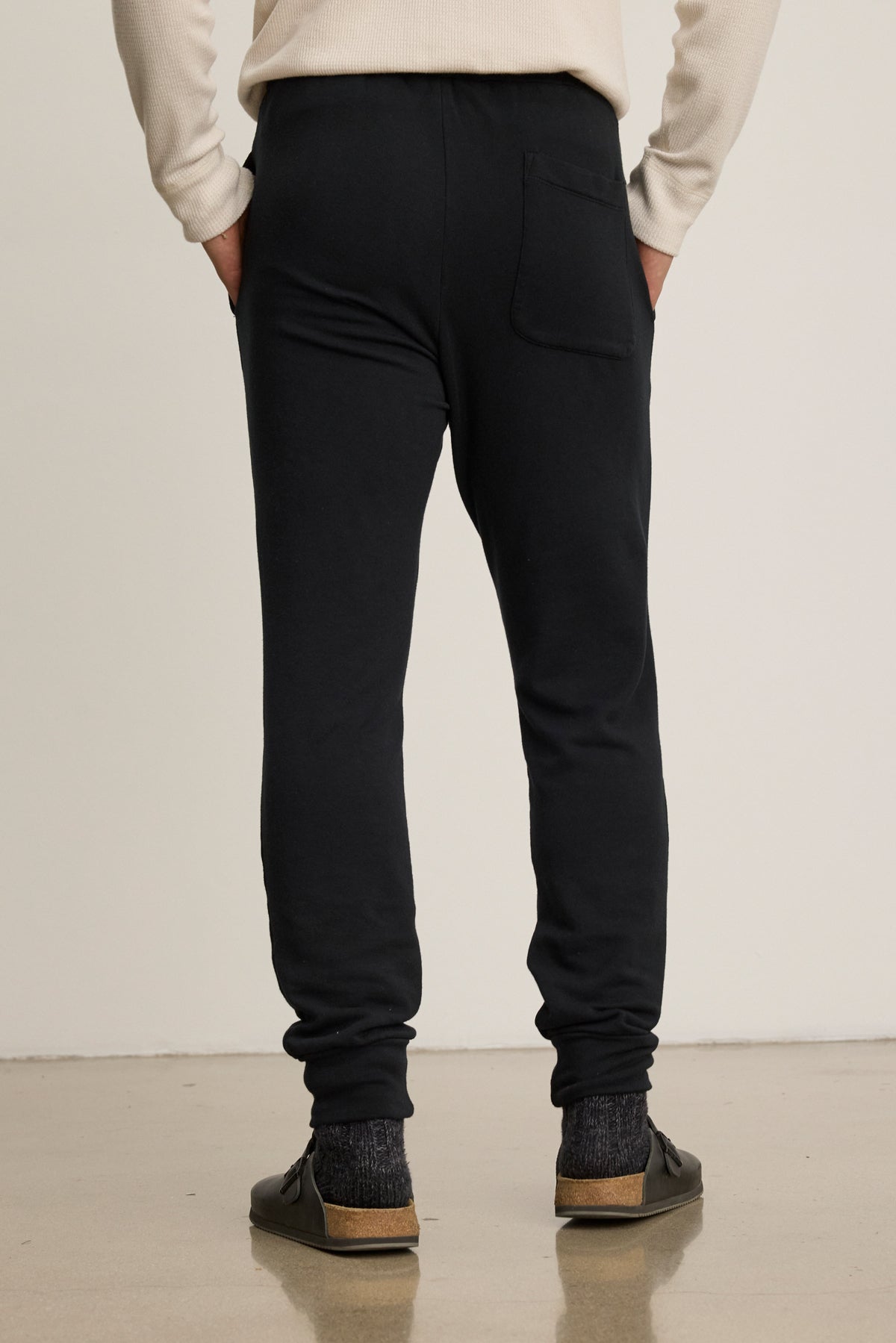 Person stands on a smooth floor, shown from behind, wearing Velvet by Graham & Spencer CROSBY JOGGER, a cream long-sleeve shirt, dark socks, and black slip-on shoes.