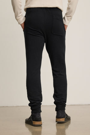 Person stands on a smooth floor, shown from behind, wearing Velvet by Graham & Spencer CROSBY JOGGER, a cream long-sleeve shirt, dark socks, and black slip-on shoes.