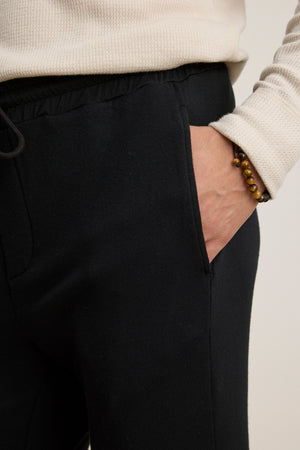 Person wearing the Velvet by Graham & Spencer CROSBY JOGGER in black with a cream ribbed sweater, one hand in their pocket, and a beaded bracelet.