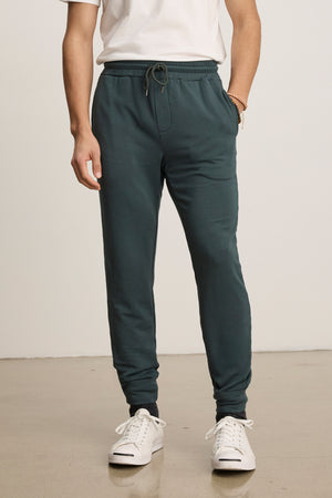 A person stands on a light-colored floor wearing a white t-shirt, white sneakers, and Velvet by Graham & Spencer’s CROSBY JOGGER—tailored joggers in dark green with a drawstring waist.