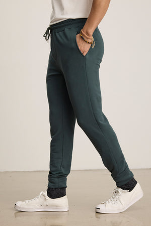 Someone stands against a plain background wearing Velvet by Graham & Spencer's CROSBY JOGGER in dark green, paired with a white t-shirt, white sneakers, and black socks.