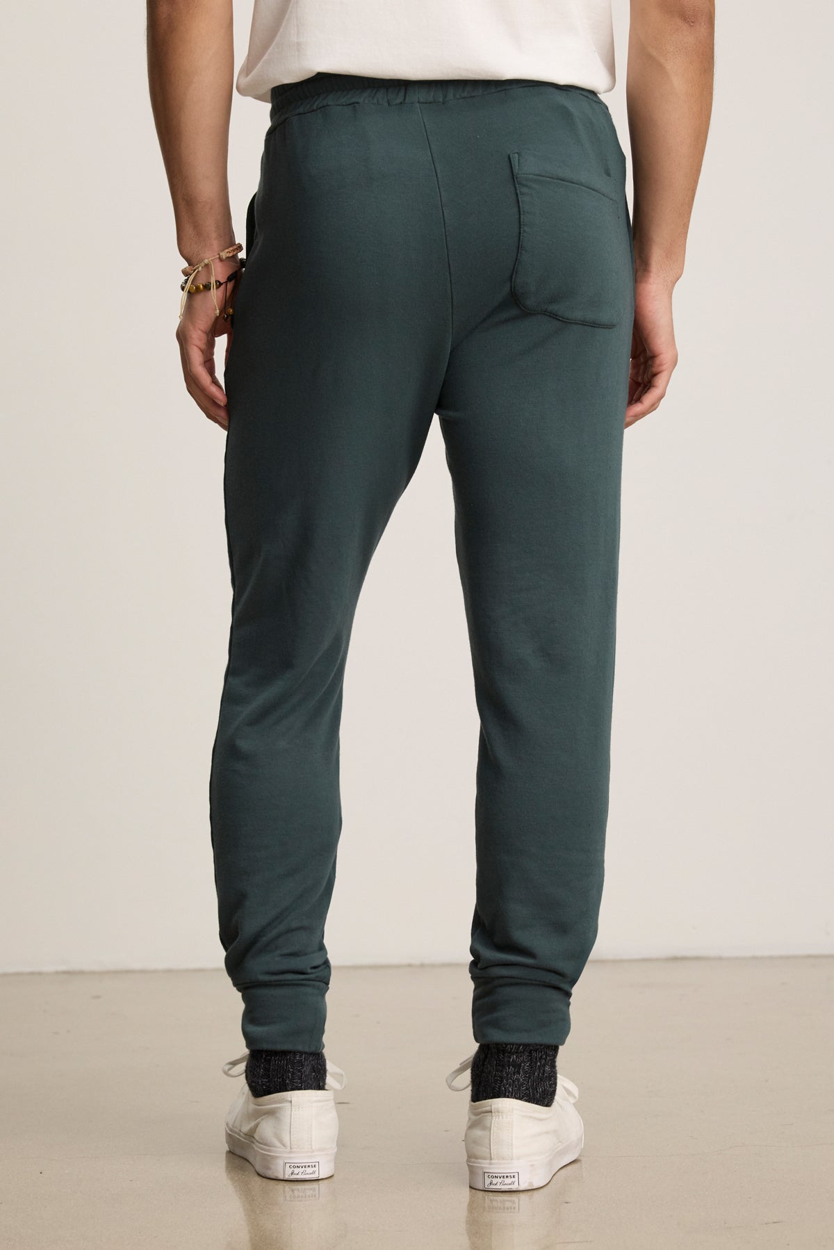 Person wearing Velvet by Graham & Spencer CROSBY JOGGER in dark green, paired with white sneakers and a white t-shirt, shown from the back standing on a light-colored floor.