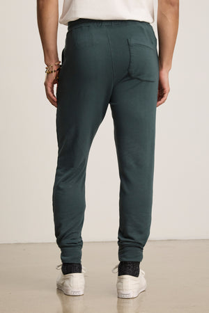 Person wearing Velvet by Graham & Spencer CROSBY JOGGER in dark green, paired with white sneakers and a white t-shirt, shown from the back standing on a light-colored floor.