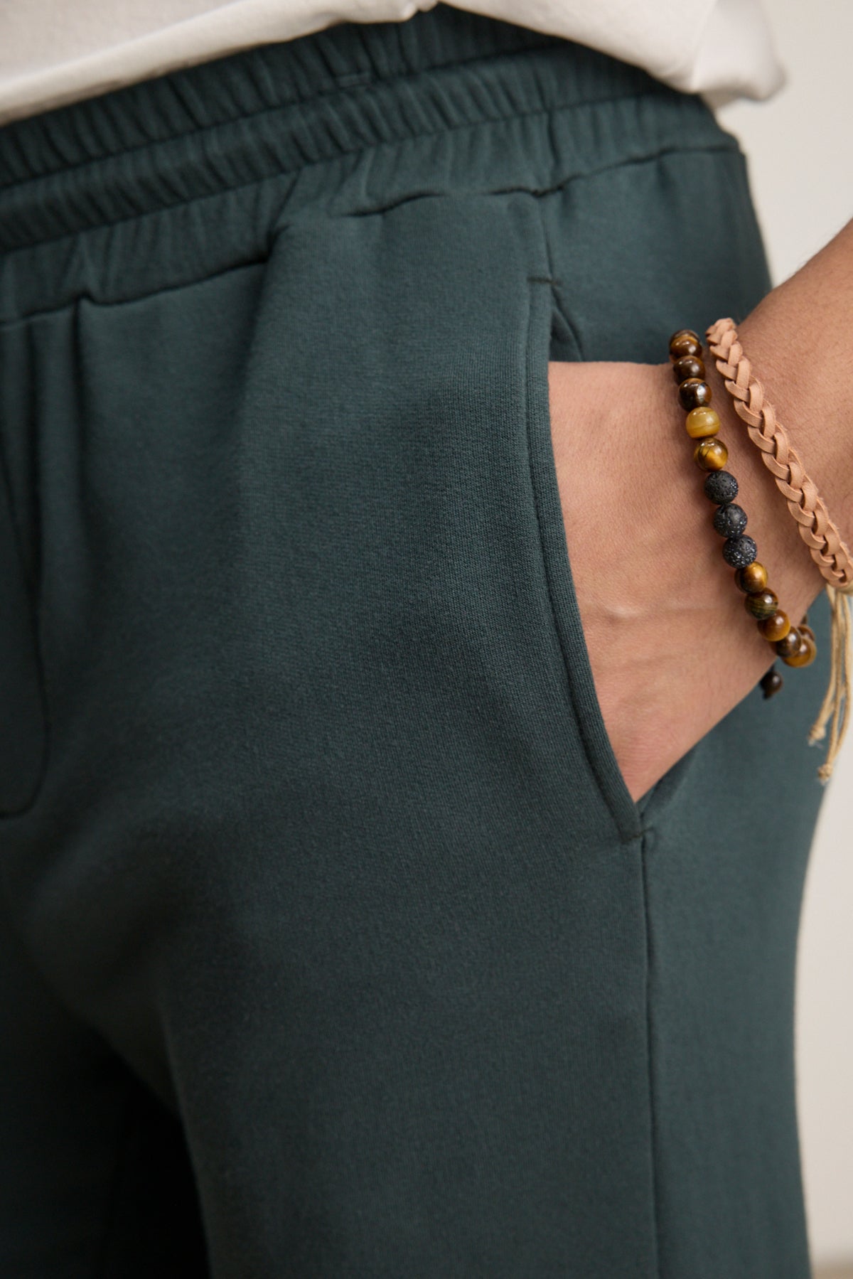A person wears the Velvet by Graham & Spencer CROSBY JOGGER in dark green, with their right hand in the pocket and beaded, braided bracelets on their wrist.