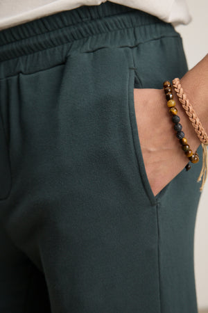 A person wears the Velvet by Graham & Spencer CROSBY JOGGER in dark green, with their right hand in the pocket and beaded, braided bracelets on their wrist.