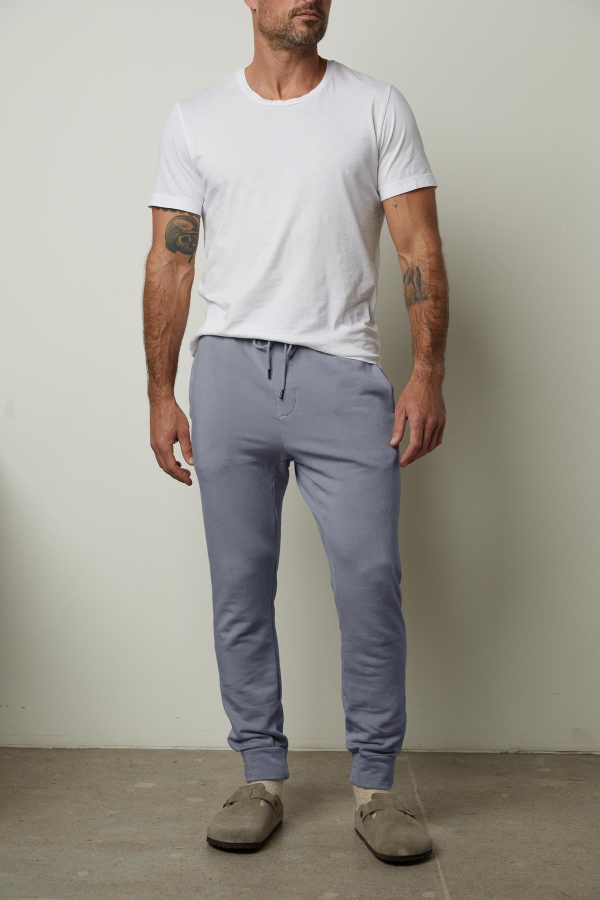 A person wearing a white t-shirt and the CROSBY LUXE FLEECE JOGGER from Velvet by Graham & Spencer stands against a plain wall. They are paired with grey slippers.