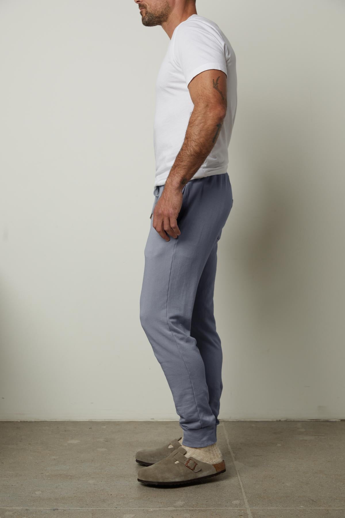 A person dressed in a white t-shirt, the CROSBY LUXE FLEECE JOGGER by Velvet by Graham & Spencer featuring a tailored design, and brown clogs stands sideways against a plain background.
