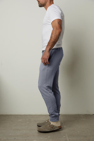 A person dressed in a white t-shirt, the CROSBY LUXE FLEECE JOGGER by Velvet by Graham & Spencer featuring a tailored design, and brown clogs stands sideways against a plain background.