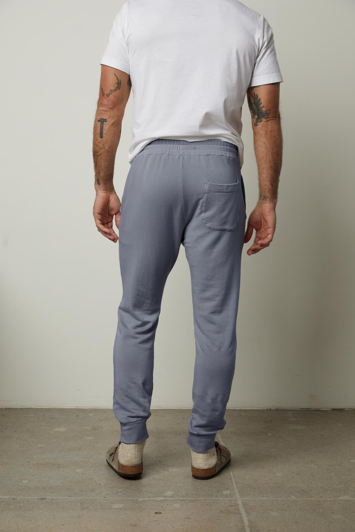 A person dressed in a white T-shirt, Velvet by Graham & Spencer's light blue CROSBY LUXE FLEECE JOGGER, and brown shoes stands facing away, with one hand in their pocket. Tattoos on their forearms suggest a dedication to workouts and customized design in everyday life.-38356001784001