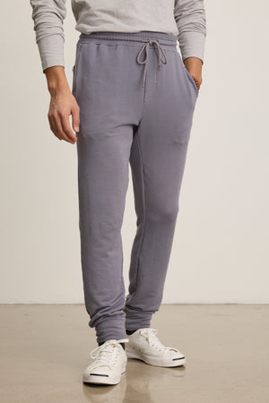 A person wearing the CROSBY JOGGER by Velvet by Graham & Spencer, paired with a light gray long-sleeve shirt and white sneakers, stands on a polished floor against a plain background.