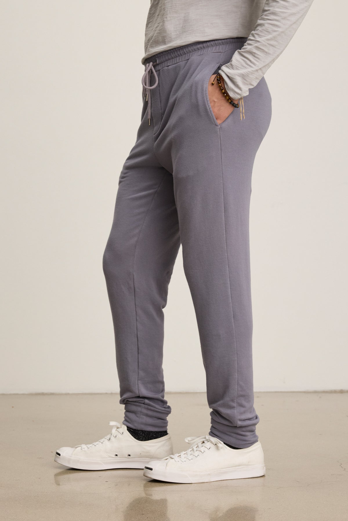 A person wears the Velvet by Graham & Spencer CROSBY JOGGER in light gray with a long-sleeve shirt, white sneakers, and bracelets, standing on a polished floor against a plain background.