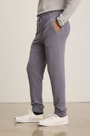 A person wears the Velvet by Graham & Spencer CROSBY JOGGER in light gray with a long-sleeve shirt, white sneakers, and bracelets, standing on a polished floor against a plain background.