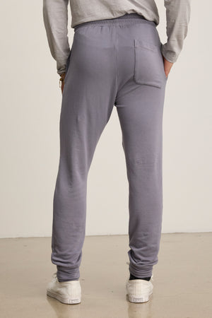 A person in light gray Velvet by Graham & Spencer CROSBY JOGGER fleece pants and white sneakers, shown from the back, stands on a smooth floor against a plain background.