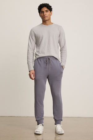 A man stands against a plain background wearing a light gray long-sleeve shirt, Velvet by Graham & Spencer CROSBY JOGGER in gray, and white sneakers.
