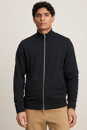 A person wears the Velvet by Graham & Spencer DEMITRI FULL ZIP black fleece jacket with front pouch pockets and tan pants, standing facing forward against a plain light background.
