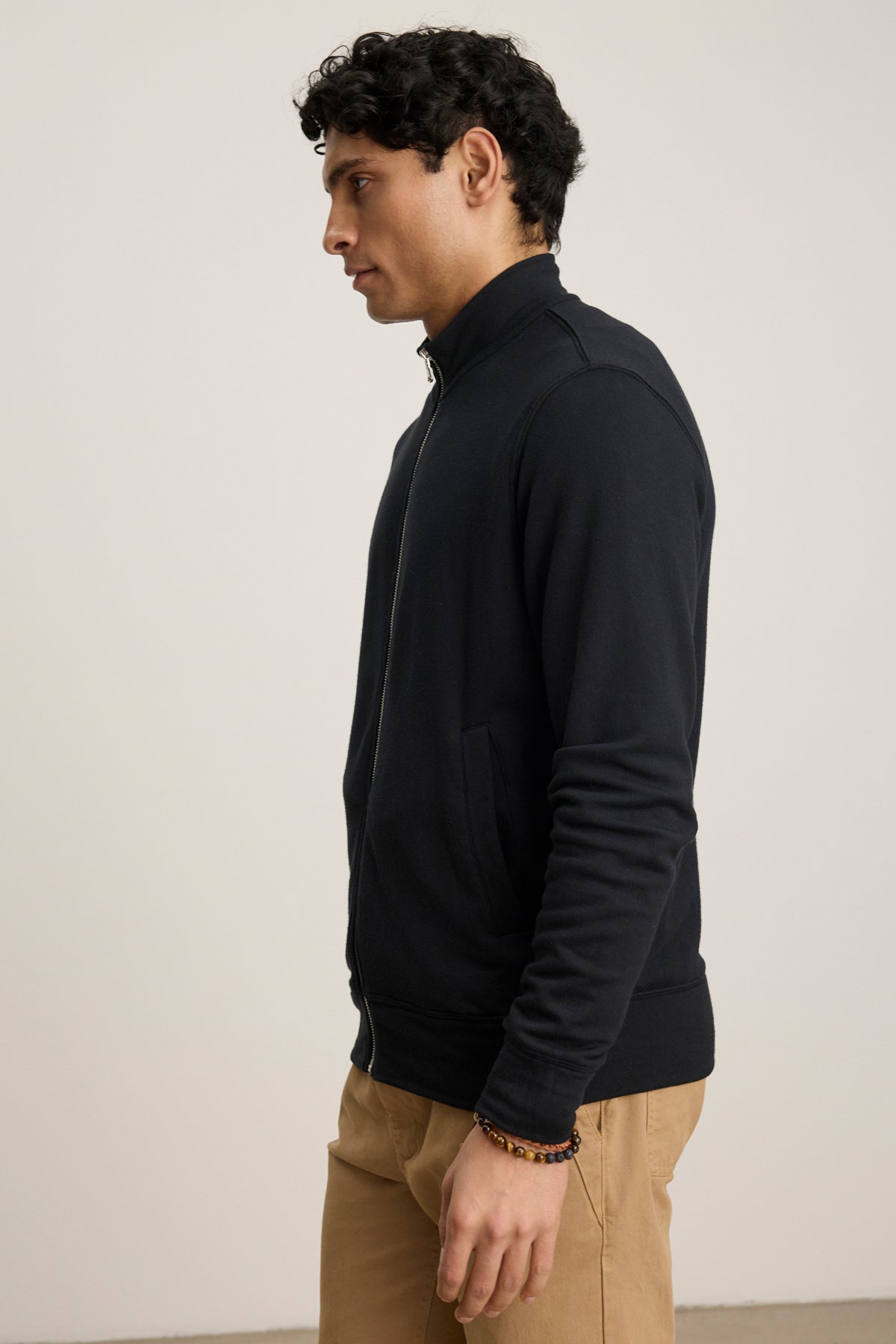 A man with short curly hair stands in profile against a plain light background, wearing the Velvet by Graham & Spencer DEMITRI FULL ZIP black fleece with front pouch pockets and khaki pants.