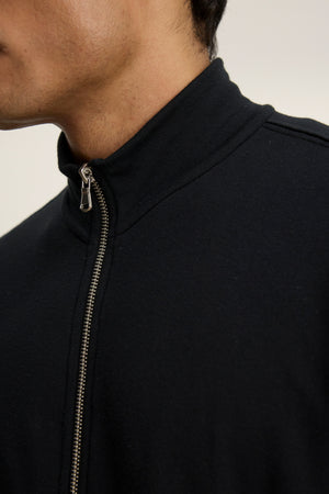Close-up of a person wearing the DEMITRI FULL ZIP black fleece jacket by Velvet by Graham & Spencer, featuring a mock neck and visible zipper; the person's face is partially out of frame.