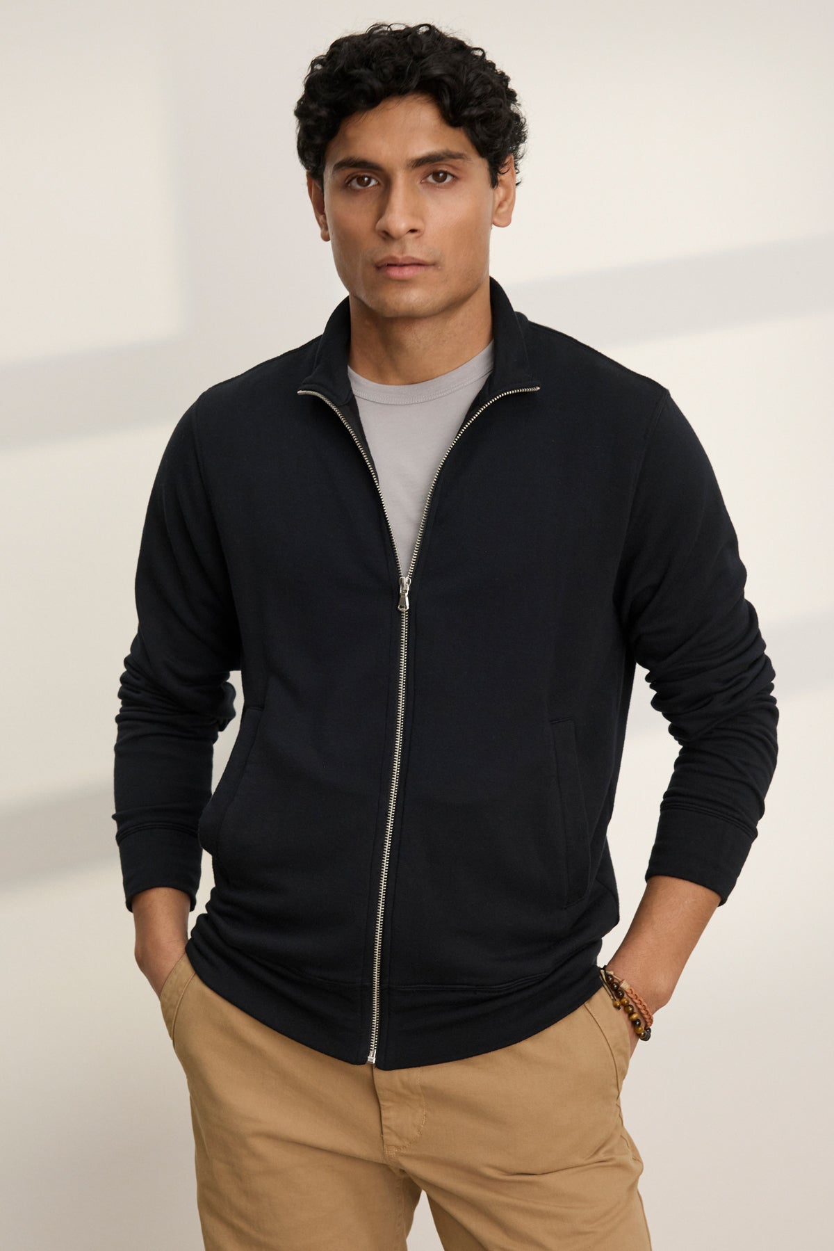 A man stands with his hands in his front pockets, wearing the Velvet by Graham & Spencer DEMITRI FULL ZIP black fleece jacket over a gray t-shirt and beige pants, set against a neutral background.