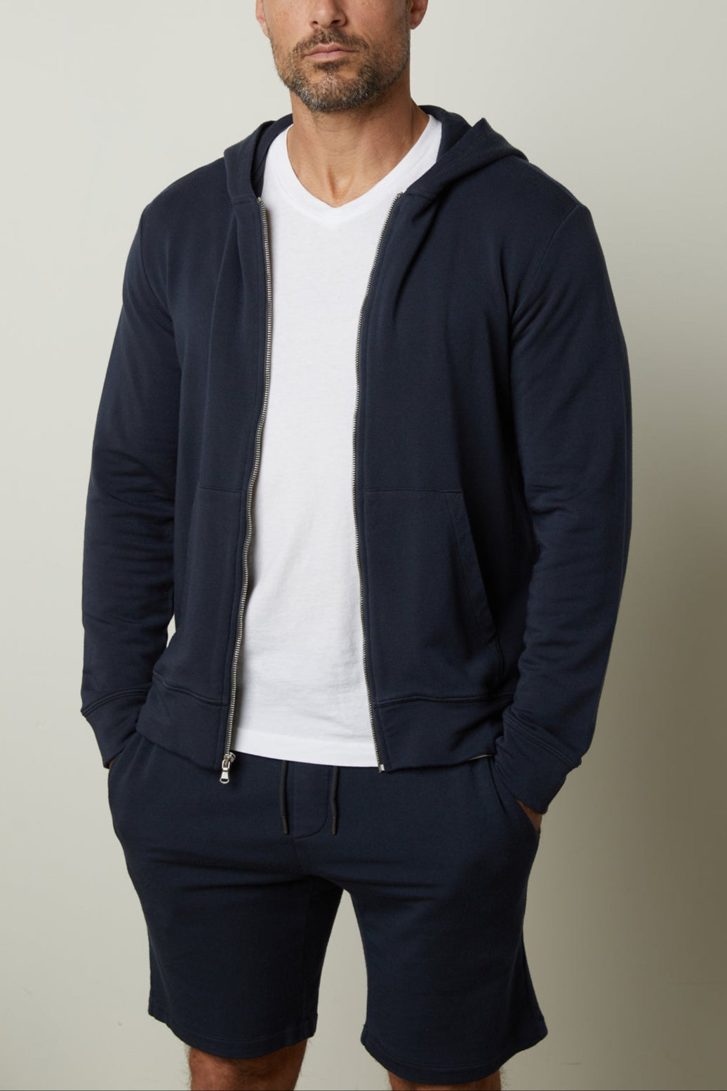 A man with a shaved head and full beard is wearing a RODAN HOODIE by Velvet by Graham & Spencer, a luxe fleece navy blue zip-up hoodie, over a white t-shirt and matching navy blue shorts, standing against a plain background.-35662630092993