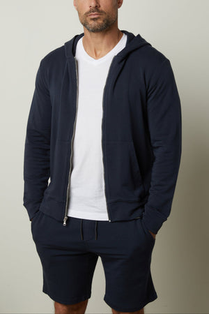 A man with a shaved head and full beard is wearing a RODAN HOODIE by Velvet by Graham & Spencer, a luxe fleece navy blue zip-up hoodie, over a white t-shirt and matching navy blue shorts, standing against a plain background.