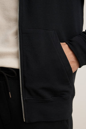 Wearing the RODAN HOODIE by Velvet by Graham & Spencer with black pants, a person has their right hand in the hoodie pocket and a white ribbed shirt is partially visible underneath.