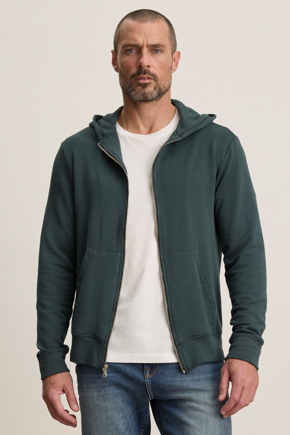 A man with short hair and a beard wears the Velvet by Graham & Spencer RODAN HOODIE in dark green luxe fleece over a white tee and blue jeans, standing against a plain light background.