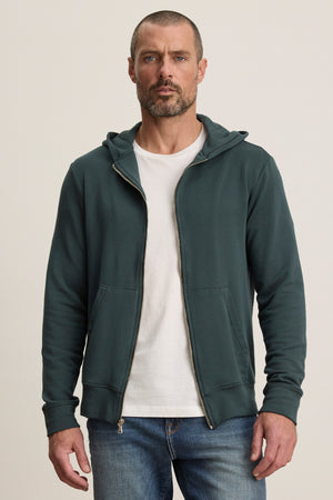 A man with short hair and a beard wears the Velvet by Graham & Spencer RODAN HOODIE in dark green luxe fleece over a white tee and blue jeans, standing against a plain light background.