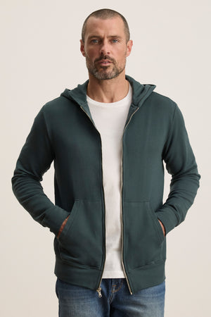 A man stands with hands in his pockets, wearing the Velvet by Graham & Spencer RODAN HOODIE over a white t-shirt and blue jeans against a plain light background.