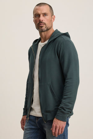 A man with short hair and a beard wears the Velvet by Graham & Spencer RODAN HOODIE, a dark green zip-up ideal as a workout layer, over a white t-shirt and blue jeans, standing against a plain light background.