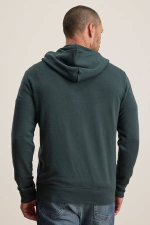 A man stands facing away from the camera in a dark green RODAN HOODIE by Velvet by Graham & Spencer, paired with blue jeans against a plain background.