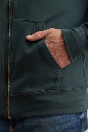 A person wearing the RODAN HOODIE by Velvet by Graham & Spencer in dark green with blue jeans has their right hand in the hoodie’s front pocket.