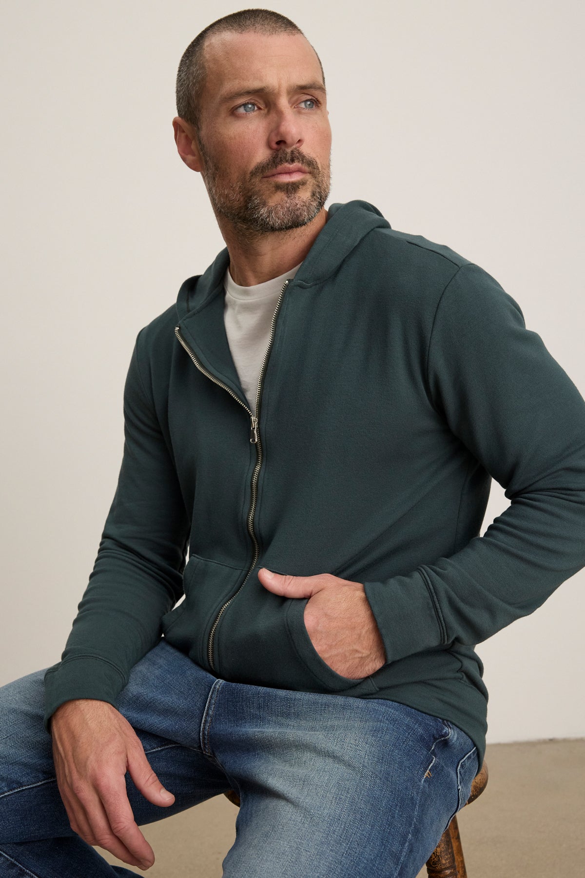 A man with short hair and a beard wears the RODAN HOODIE by Velvet by Graham & Spencer and jeans, sitting on a chair with one hand in his pocket, looking off to the side.-39905810383041