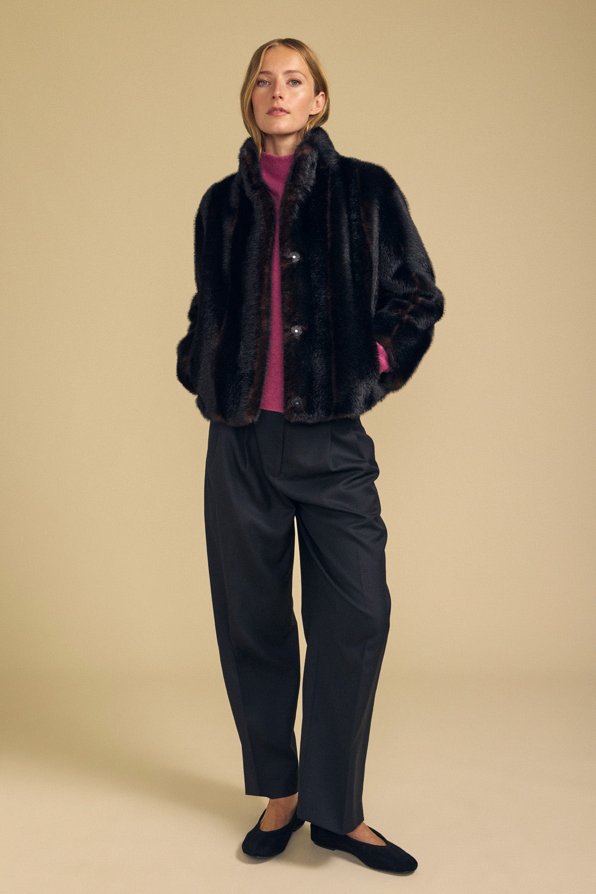 A woman stands against a beige background wearing the Velvet by Graham & Spencer SHEENA FAUX FUR JACKET, styled with a magenta sweater, loose black trousers, and black flat shoes, her hands tucked in the jacket pockets for a cozy fit.-42063845130433