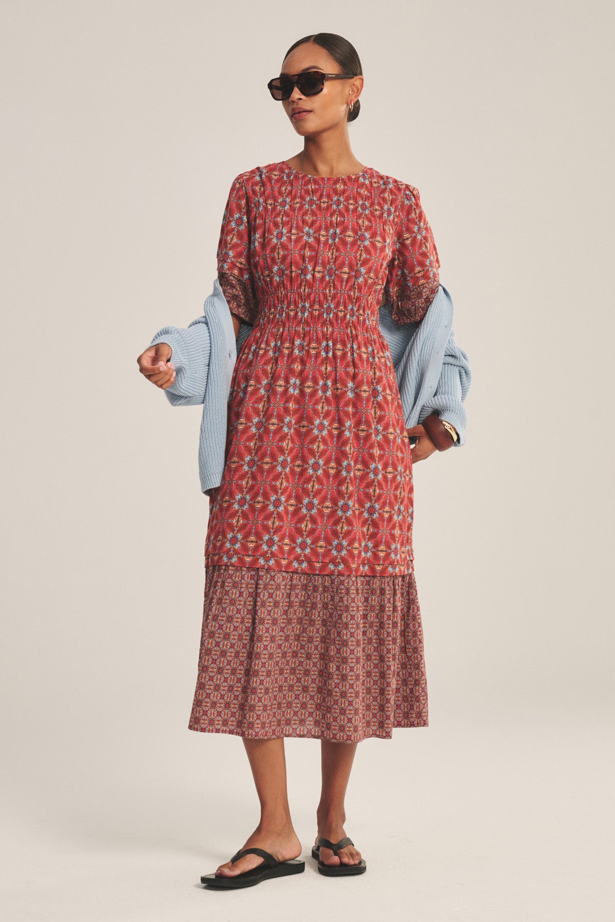 A woman in sunglasses, black sandals, and a light blue sweater draped over her shoulders wears the MAELA DRESS by Velvet by Graham & Spencer—a red smocked waist midi dress with a tiered hem—against a plain background.-41089019379905