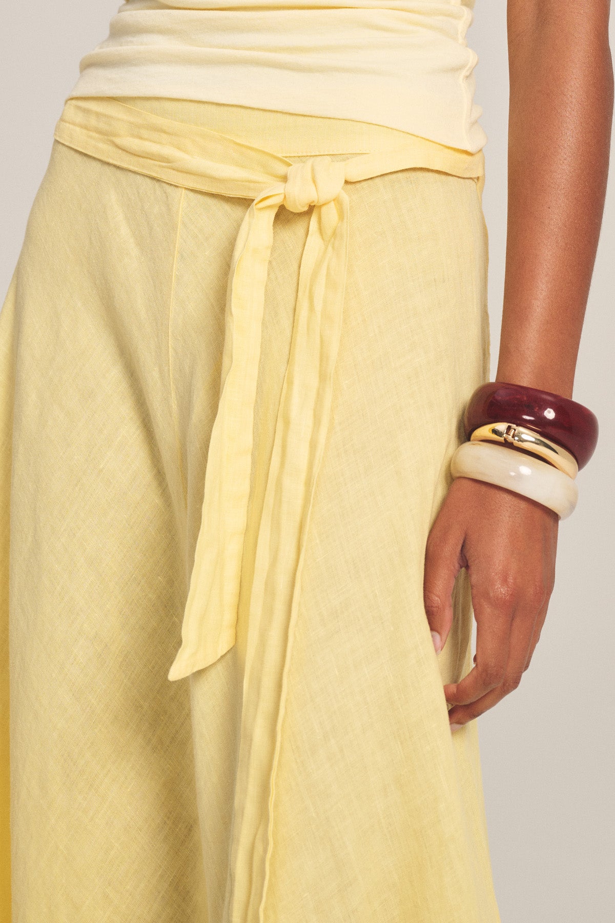 A person wears the MALINA LINEN SKIRT by Velvet by Graham & Spencer in yellow with a light yellow top, styled with three chunky bangles in white, gold, and burgundy.-41128159314113