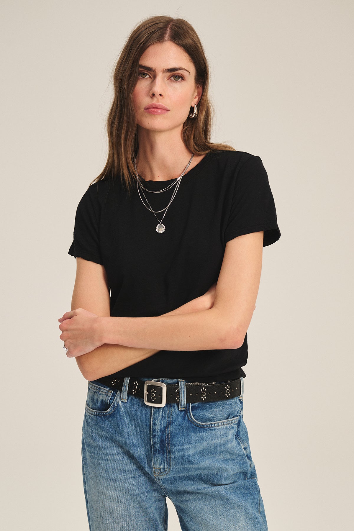 A woman with long brown hair wears the MARIE TEE by Velvet by Graham & Spencer in black cotton slub, paired with blue jeans, a black belt, and layered necklaces. She stands with arms crossed against a plain background.