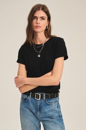 A woman with long brown hair wears the MARIE TEE by Velvet by Graham & Spencer in black cotton slub, paired with blue jeans, a black belt, and layered necklaces. She stands with arms crossed against a plain background.