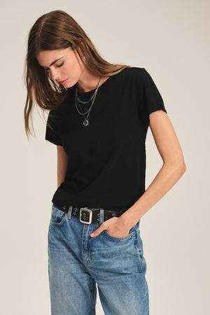 A woman wears the MARIE TEE by Velvet by Graham & Spencer, styled with blue jeans, a black belt, and layered necklaces as she stands against a plain background, looking down.