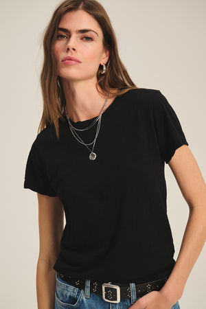 A woman with long brown hair wears the MARIE TEE by Velvet by Graham & Spencer, styled in a boxy fit with layered silver necklaces and blue jeans, standing against a plain background.