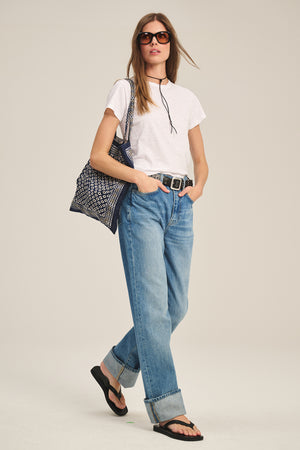A woman in sunglasses wears the MARIE TEE by Velvet by Graham & Spencer—a white cotton slub, boxy-fit casual top—paired with blue cuffed jeans, black sandals, and a patterned tote bag against a plain background.
