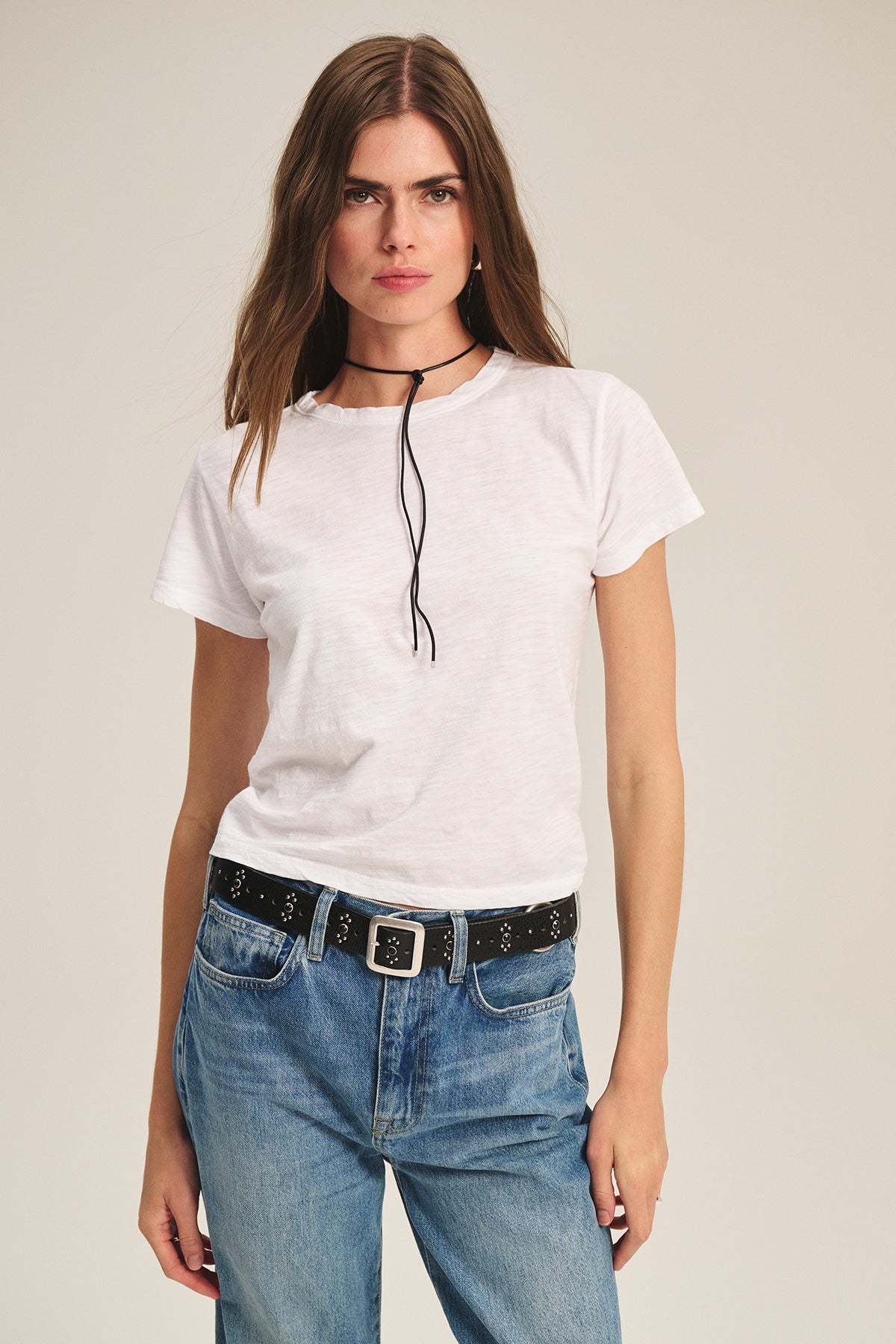 A woman stands against a plain background wearing the MARIE TEE by Velvet by Graham & Spencer, styled with blue jeans, a black studded belt, and a thin black choker.