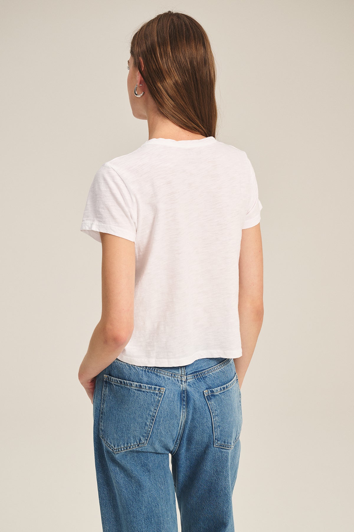 A person stands with their back to the camera, wearing the MARIE TEE by Velvet by Graham & Spencer—a white cotton slub casual tee—paired with blue jeans against a neutral background.