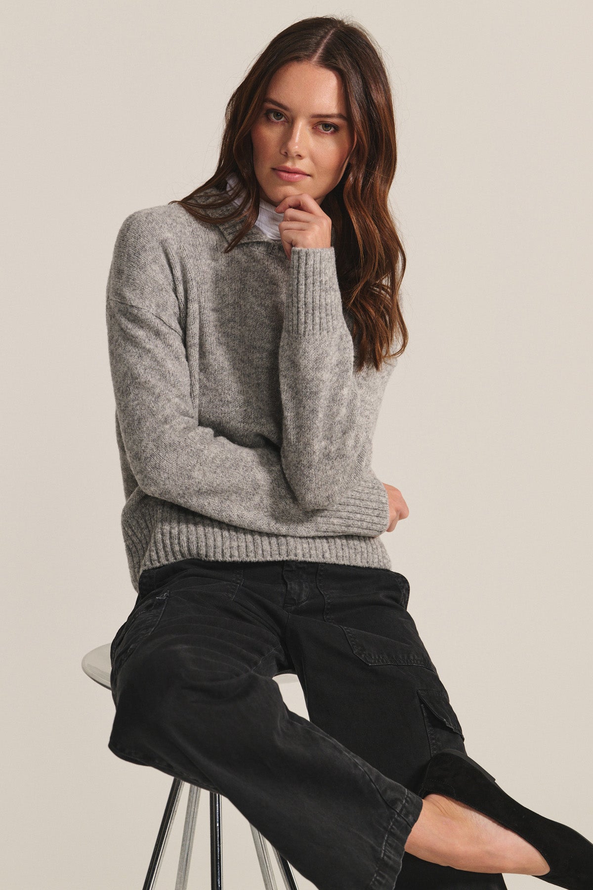A woman with long brown hair wears the BRIELLE SWEATER by Velvet by Graham & Spencer and black pants while sitting on a stool, resting her chin on her hand against a plain background.-40333255344321