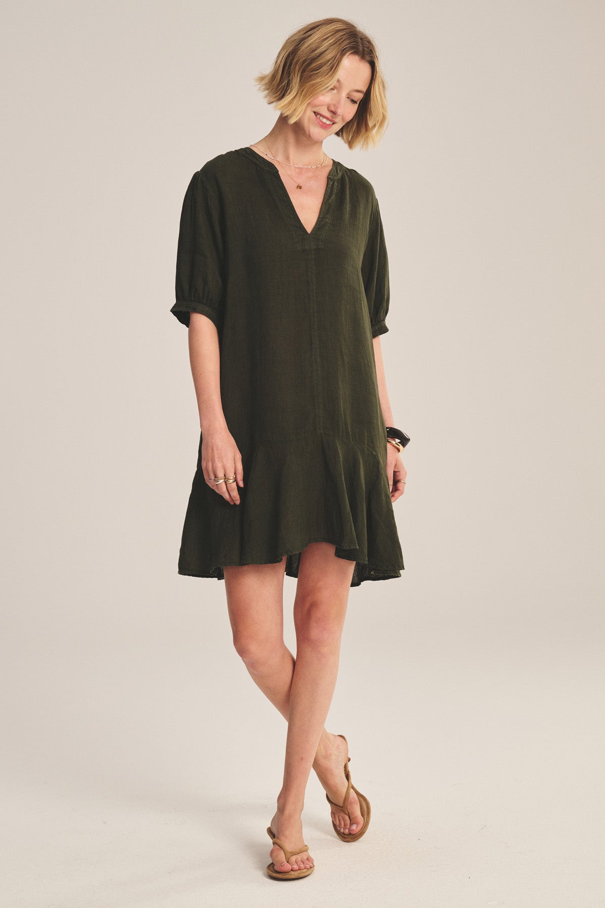 A woman wears the MEADOW DRESS by Velvet by Graham & Spencer—a loose, dark green linen dress with short sleeves, V-neckline, and ruffle-tiered hem—in a studio setting, paired with flat sandals.-41338482524353